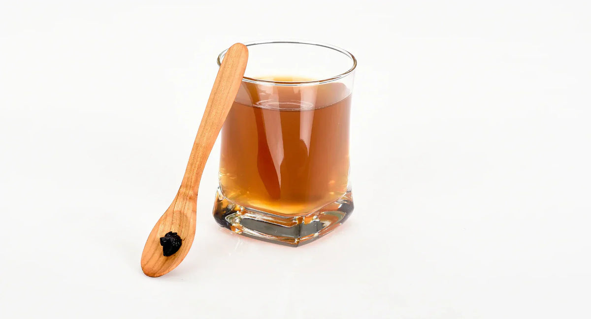 Glas mit einer bräunlichen Flüssigkeit und einem Löffel mit Shilajit-Harz daneben